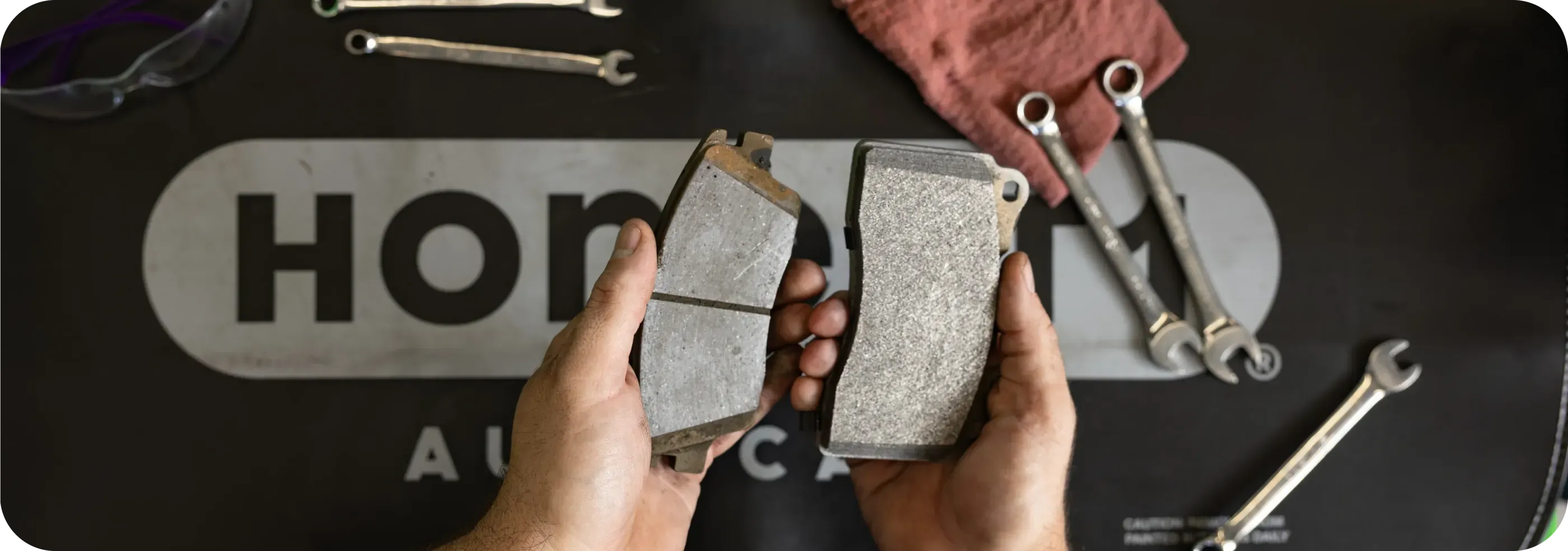 Worn and new brake pads held over an Honest1 Auto Care workbench